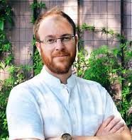 Man with glasses and red beard, arms crossed, smiling. White shirt, outdoors, greenery background.