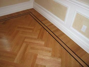 Corner of a wood floor with herringbone pattern and dark border trim beside beige walls.