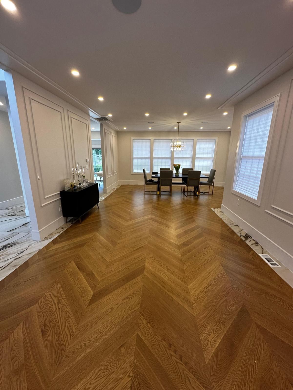 Bright empty dining room with herringbone wood floor, recessed lights, and large windows at the far end