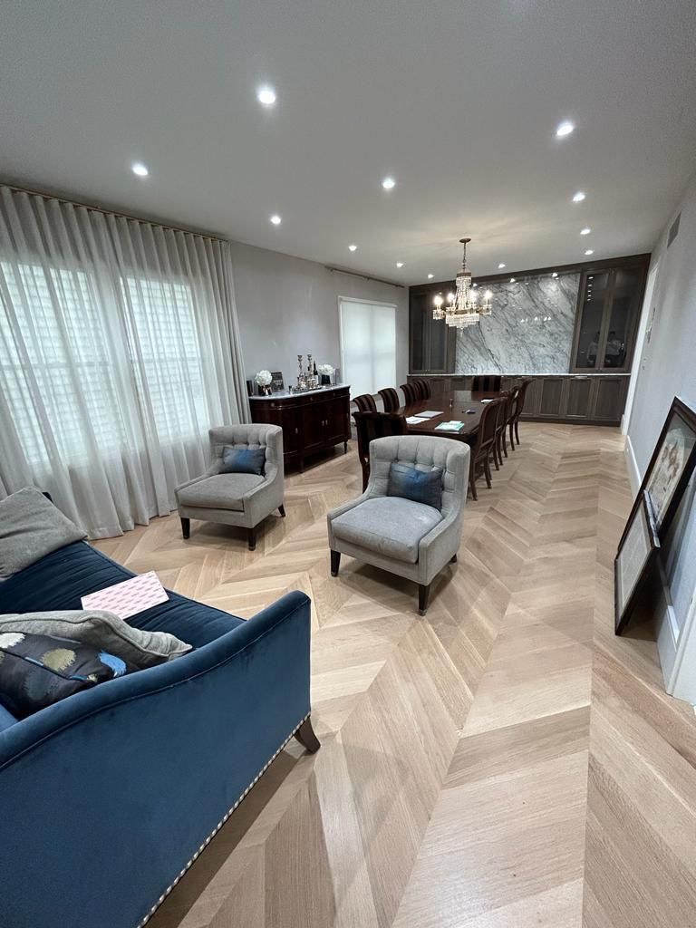 Modern living room with blue chairs, dining table, herringbone floor, and large curtained windows