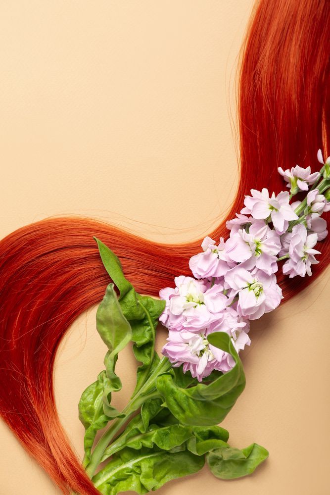 I capelli rossi scorrono su uno sfondo neutro, intrecciati con un rametto di fiori viola chiaro e foglie verdi.