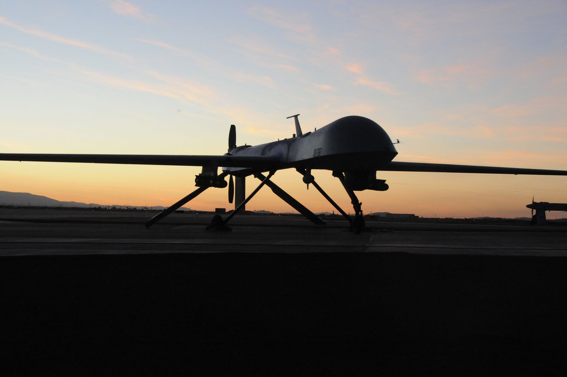 A silhouette of a military drone parked on a tarmac at sunset with a pastel sky in the background.