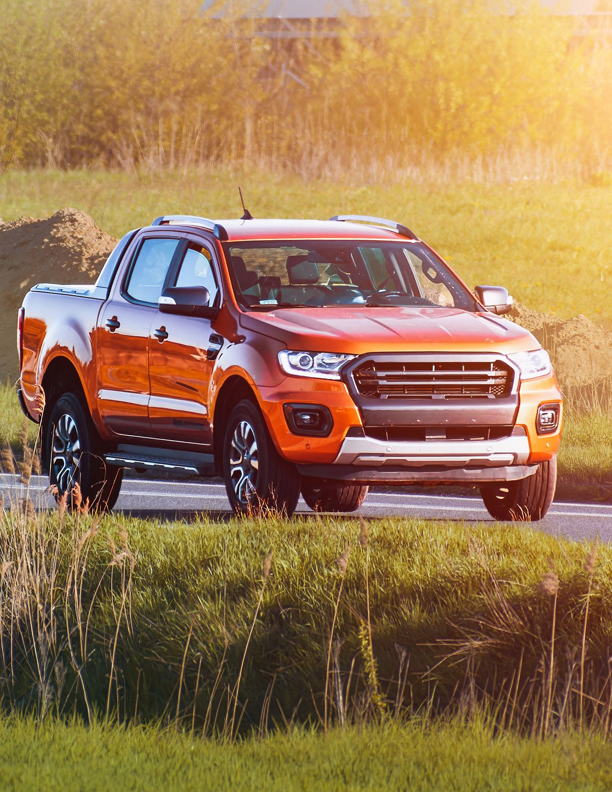 An orange pickup truck driving on a paved road through a grassy field at sunset.