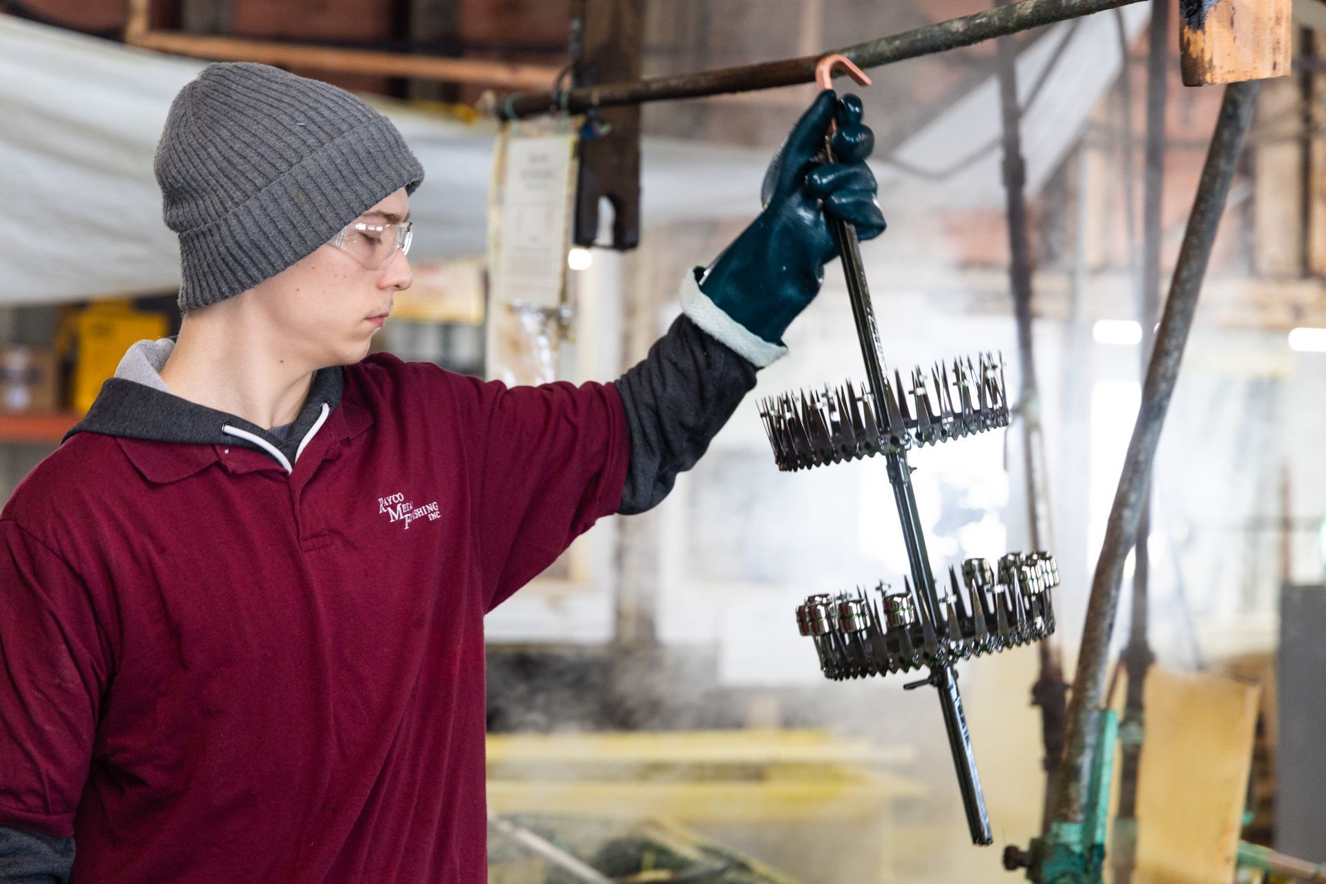 A person wearing a beanie and protective glove holds a rack of small metal parts over steam in a workshop.