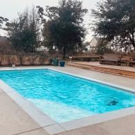 A bright blue rectangular swimming pool with concrete decking, surrounded by trees and outdoor furniture on a cloudy day.