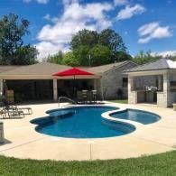 A backyard swimming pool with a kidney shape next to a covered patio and a stone outdoor kitchen under a roof.