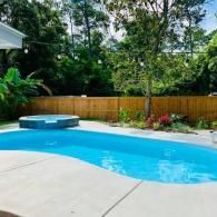A kidney-shaped swimming pool with a small hot tub sits in a backyard with a wooden fence and lush green trees.