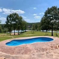 A bright blue, kidney-shaped swimming pool sits on a stamped concrete patio, overlooking a lake and grassy trees.