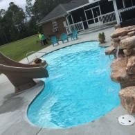 A backyard swimming pool with a curved slide, rock water feature, and patio seating near a house.