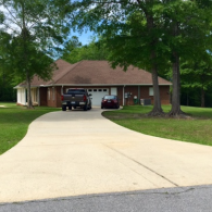 A wide concrete driveway leads to a brick house with a brown roof and a two-car garage, framed by several large trees.