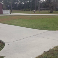 A concrete driveway curves through a front yard with grass toward a white fence and a tall flagpole.