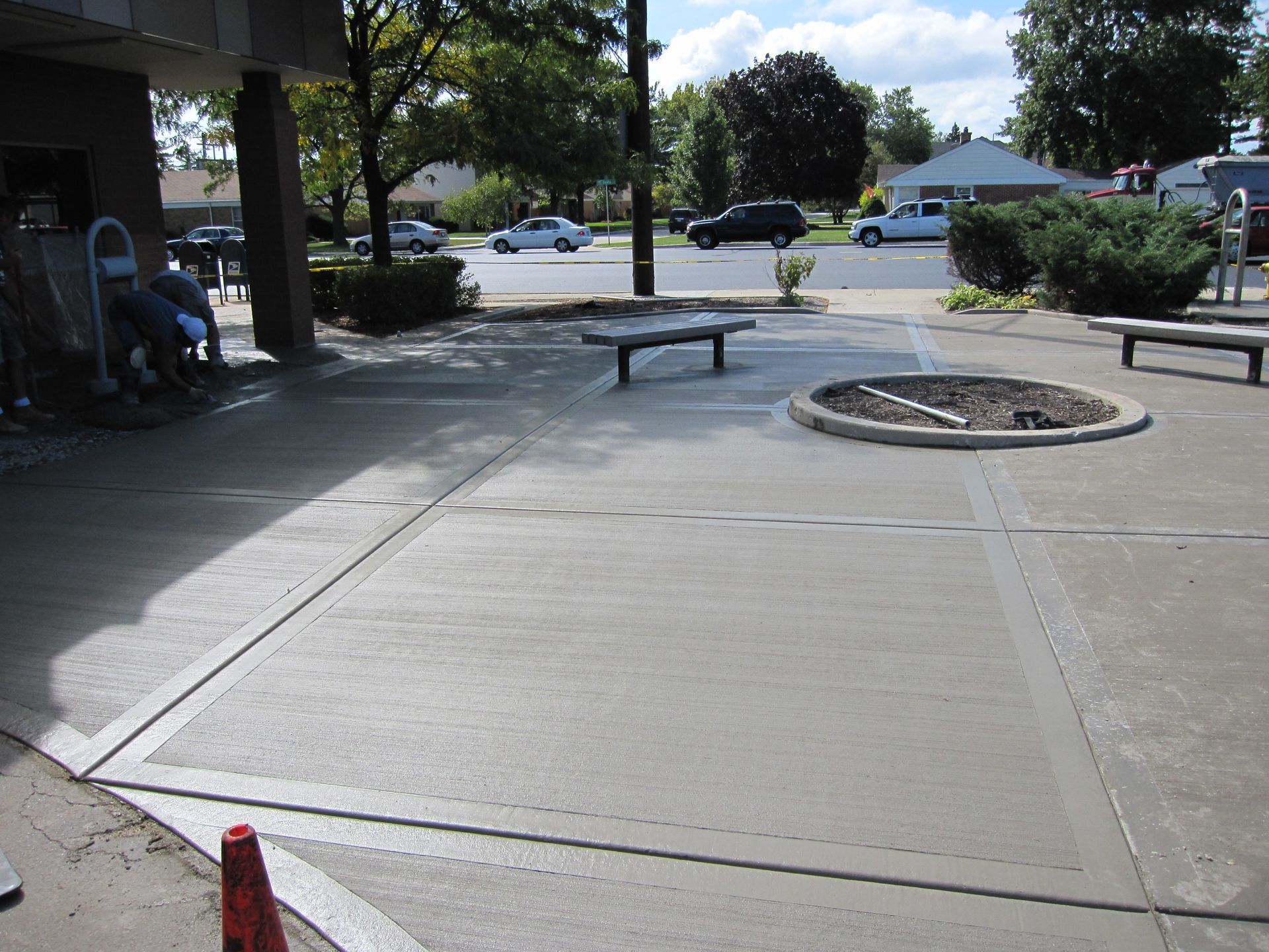 Concrete patio with benches, fire pit, and people seated near a building entrance on a sunny day.