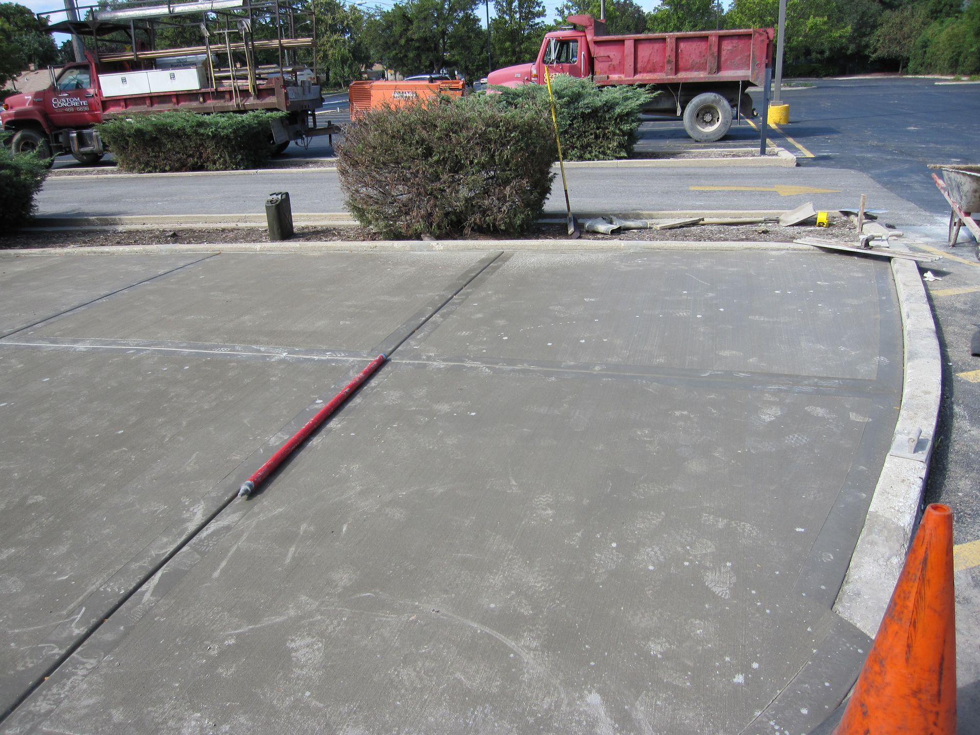 Freshly poured concrete in a parking lot, with trucks and bushes in the background, and an orange safety cone in the foreground.