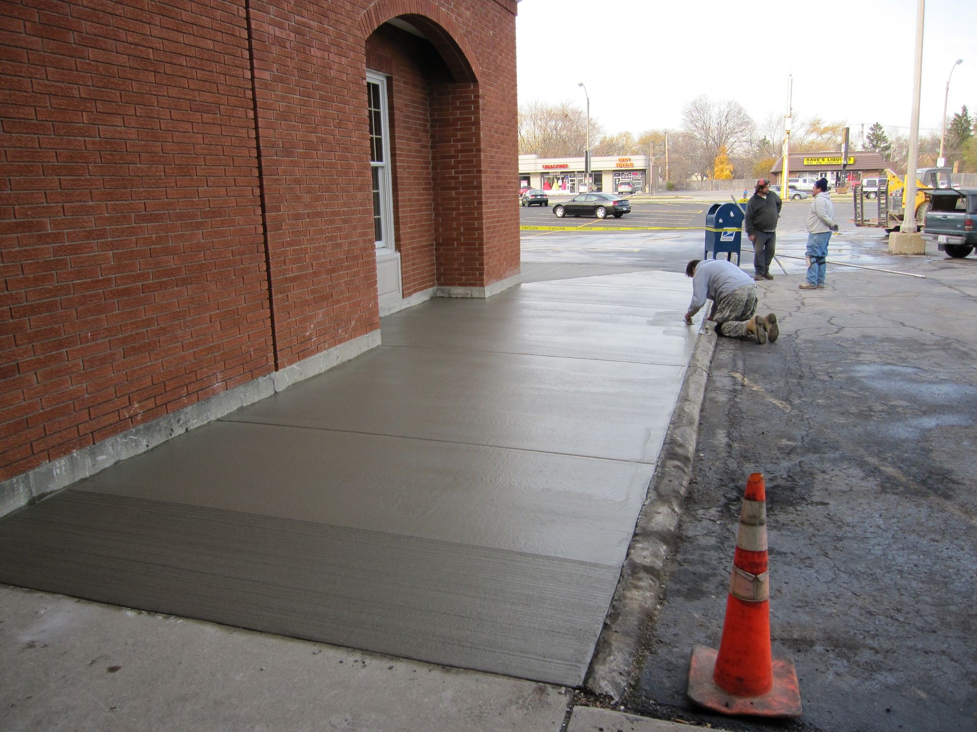 Workers finishing a concrete sidewalk by a brick building, with an orange traffic cone in front.