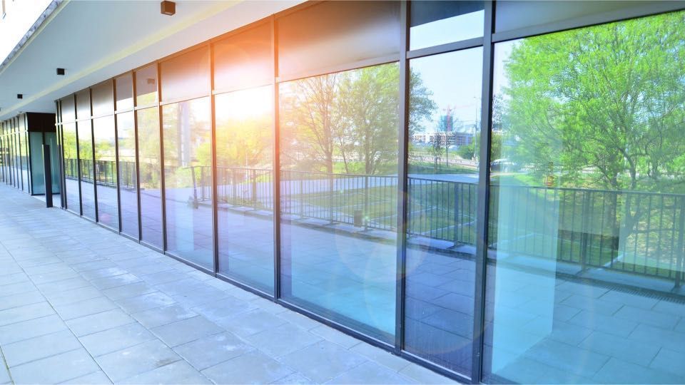 Exterior view of a modern building with a long row of large glass windows reflecting trees and sky; sunlight shining.