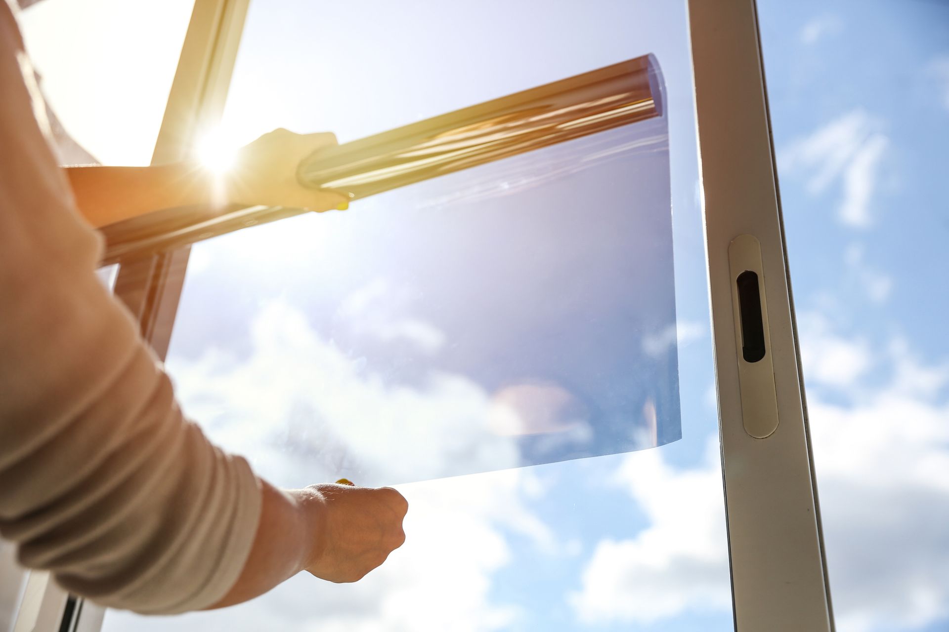 Person installing tinted film on a window with a bright sky in the background. Hands hold the film and a tool to apply it.