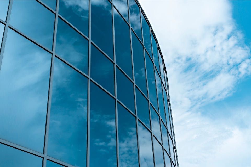Glass facade of a modern building reflecting a bright blue sky with fluffy white clouds.