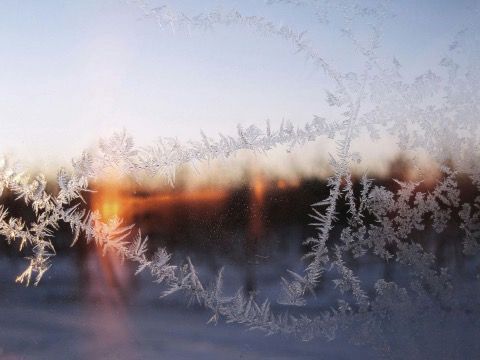 Frosty window with intricate ice crystals, obscuring a winter sunset in shades of orange and yellow.
