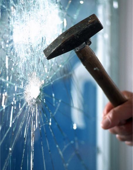 A hand holds a hammer striking a window, creating a large hole and spreading cracks.
