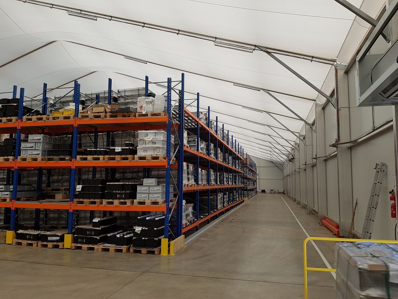 Longue allée d'entrepôt avec de hautes étagères de stockage bleues et oranges sous un plafond blanc en forme de tente.