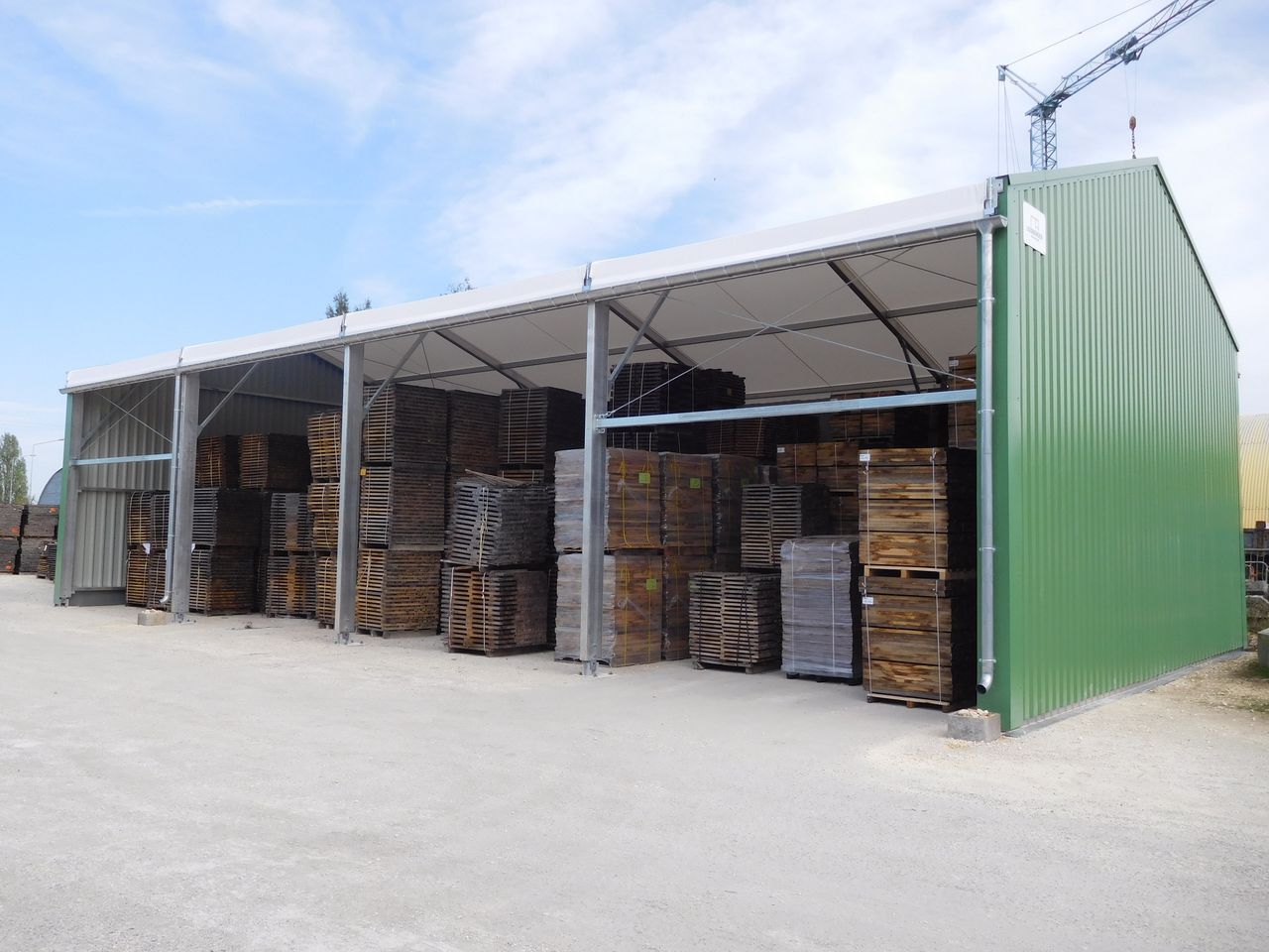 Abri métallique vert ouvert sur les côtés, servant au stockage extérieur de barils et de palettes en bois empilés.