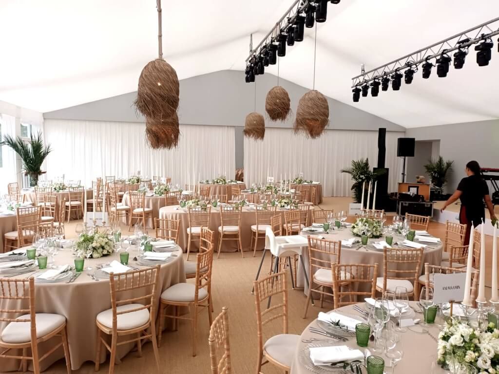 Élégante tente blanche pour réception de mariage avec tables rondes, chaises en bois et guirlandes lumineuses tissées.