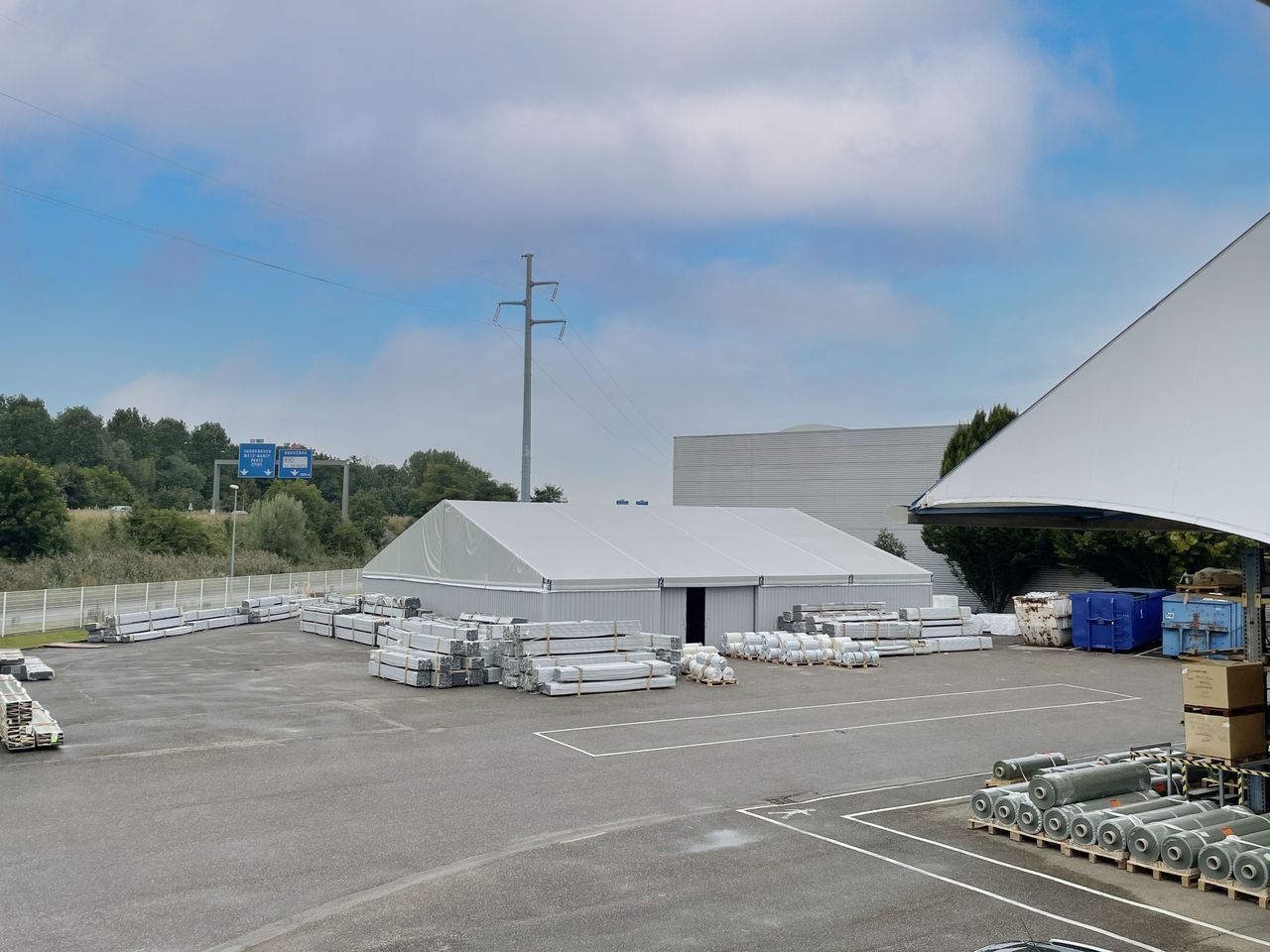Zone industrielle avec des piles de matériaux emballés, des tentes blanches et des conteneurs bleus sous un ciel nuageux.