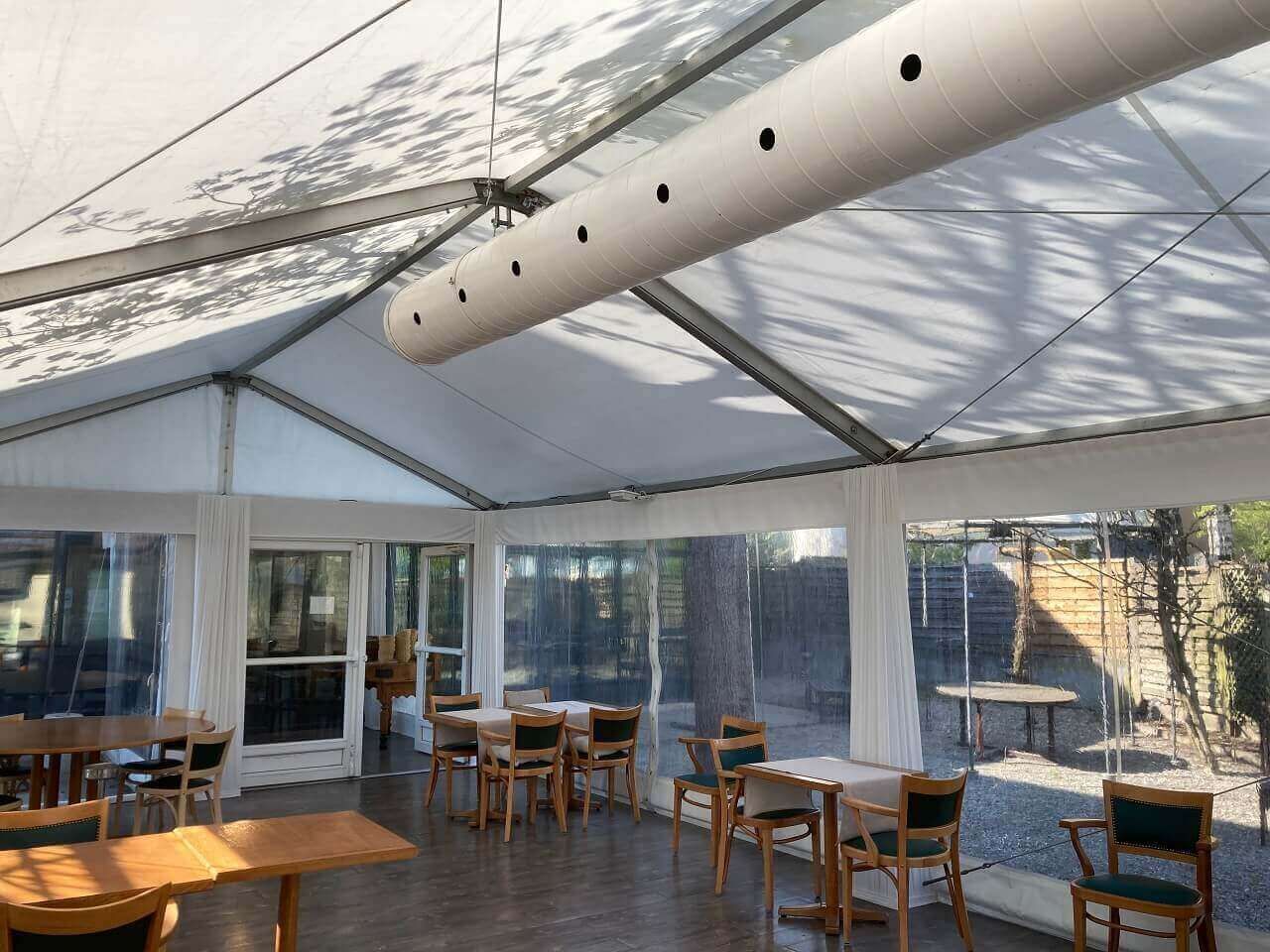 Espace repas extérieur vide sous une tente, avec tables et chaises en bois et parois en plastique transparent.