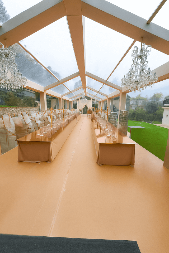 Longue tente de banquet au toit transparent, lustres et tables dressées sur un sol beige à côté d'une pelouse verte