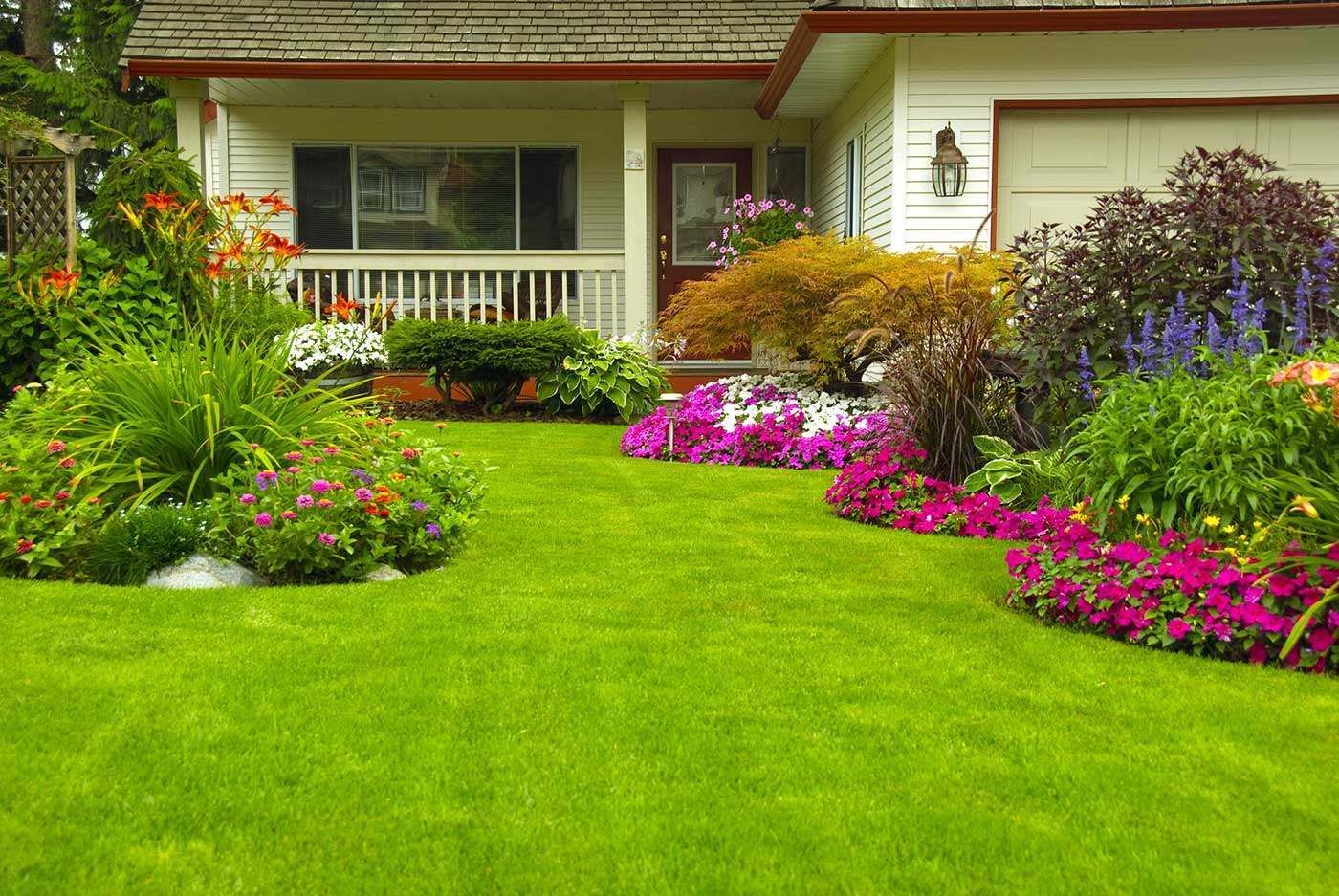 Lush green lawn with vibrant flower beds bordering a white house with a porch and garage.