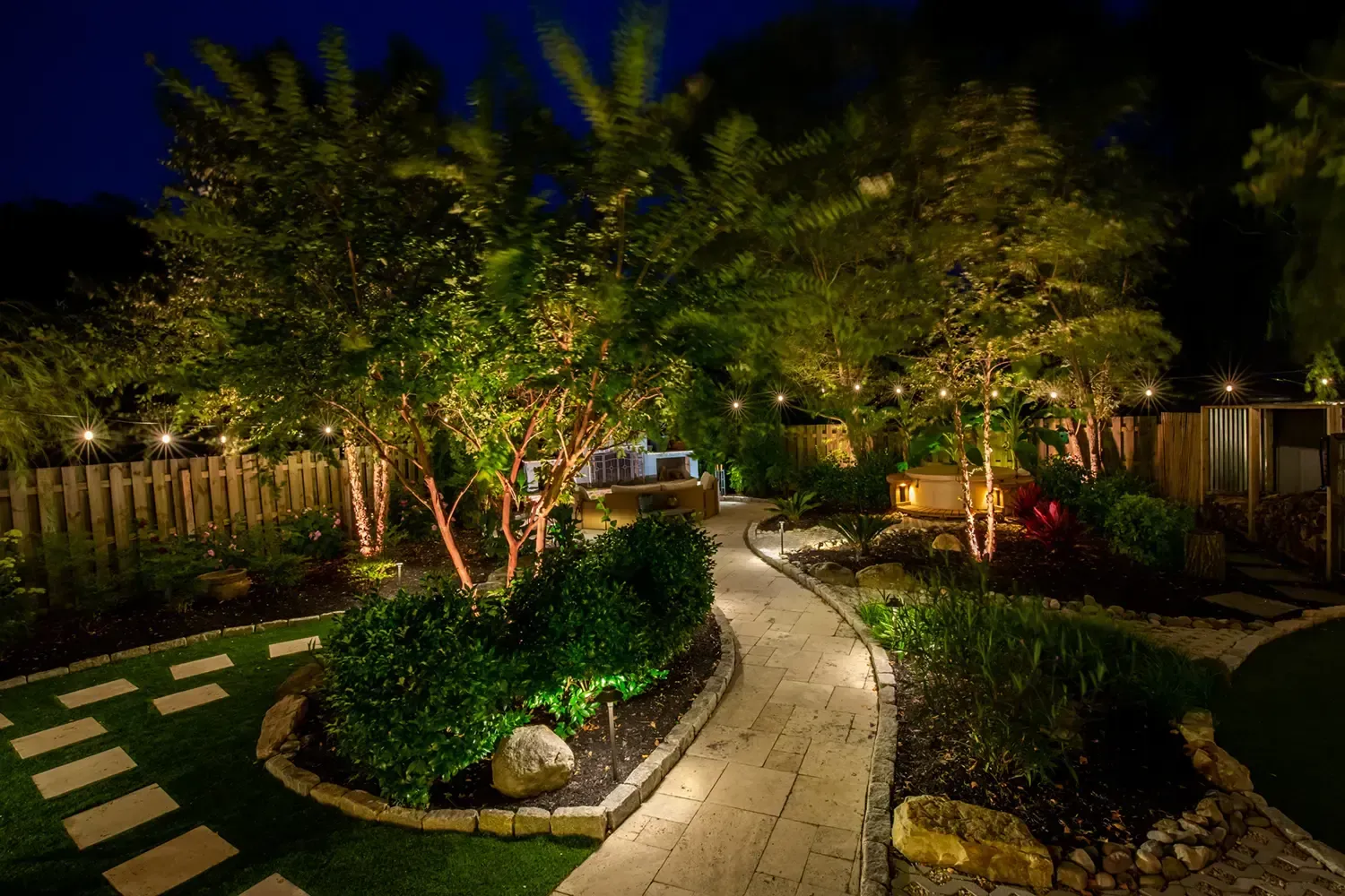 Night view of a lit garden path winding through trees with lights, a stone pathway, and lush landscaping.
