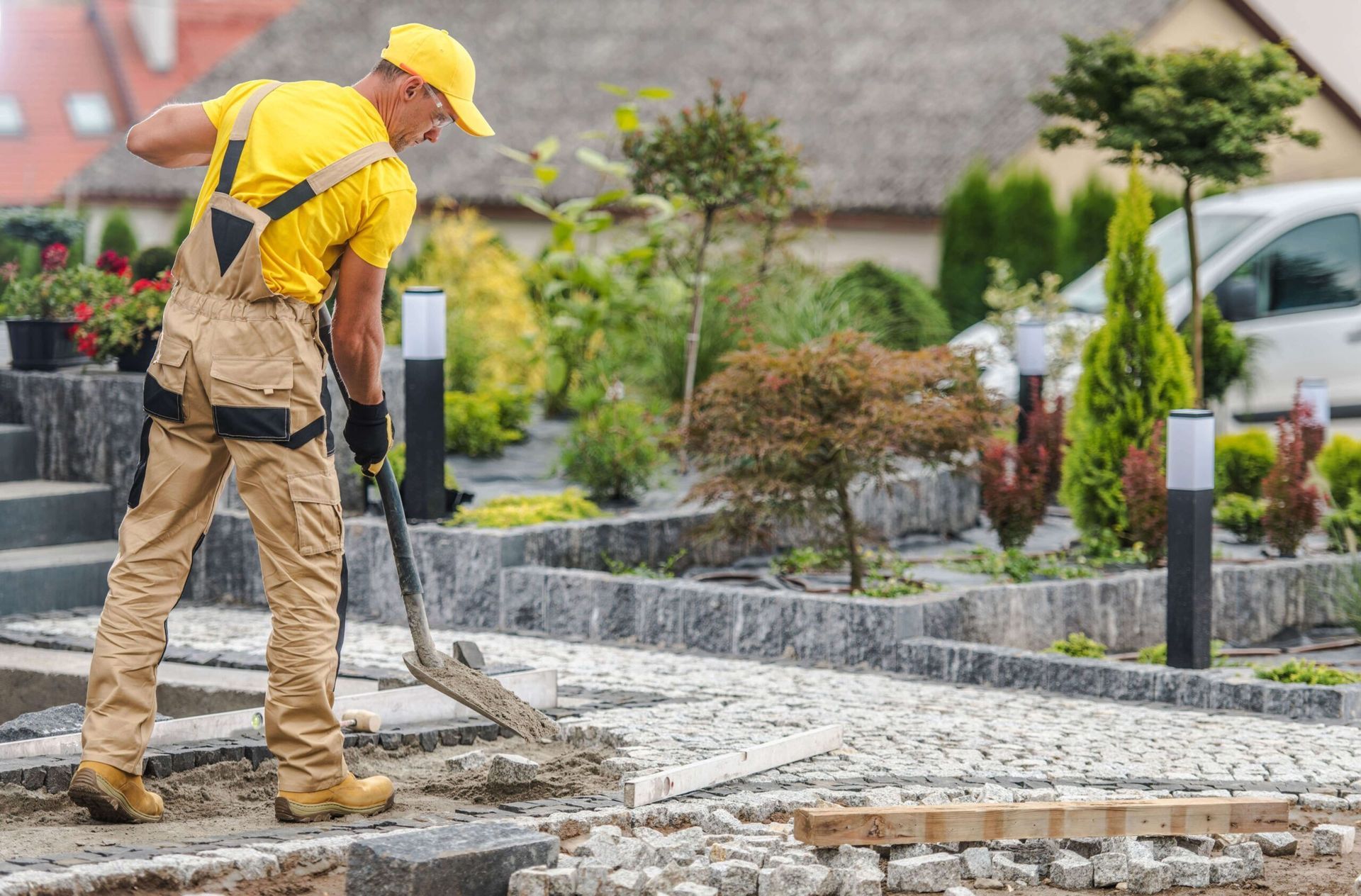 Man in yellow shirt and overalls shoveling on a paved path in a garden.