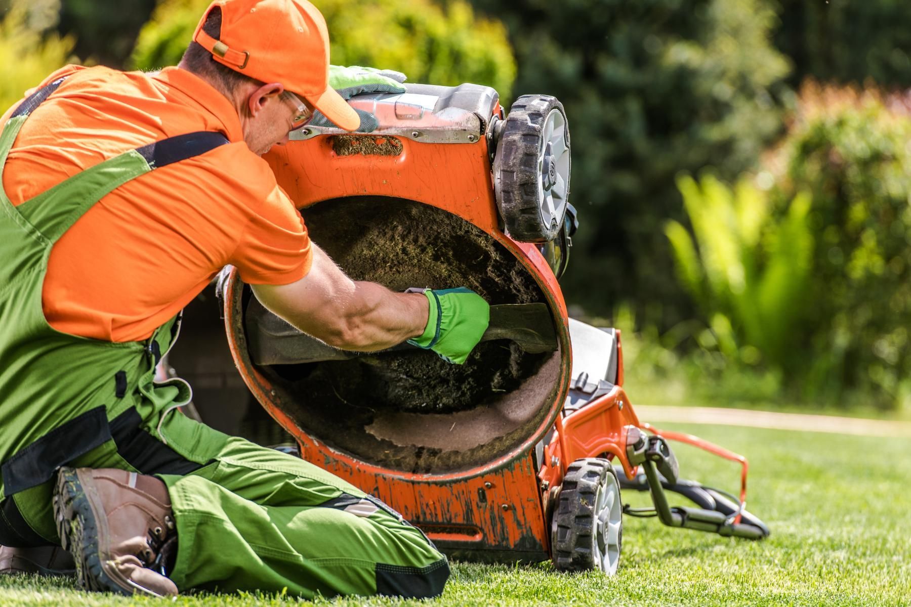 Man in orange and green work clothes, inspecting the blade of an orange lawnmower on a green lawn.