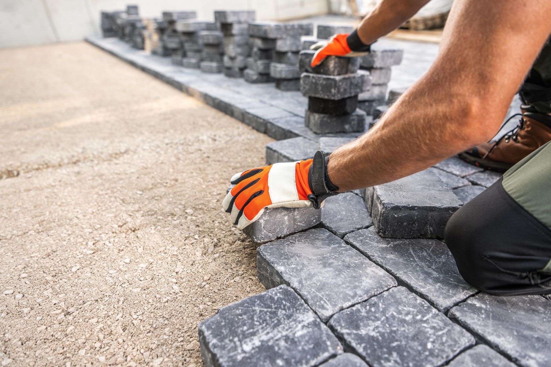 Person wearing orange gloves laying dark paving stones on a gravel base.