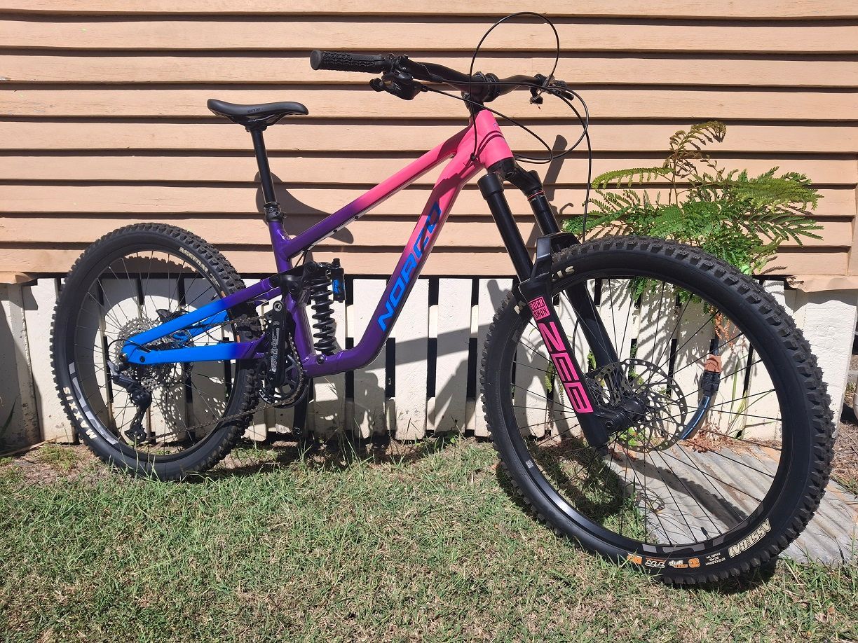 Mountain bike with pink, purple, and blue frame leaning against a wooden wall on grass — City Centre Cycles in Berserker, QLD