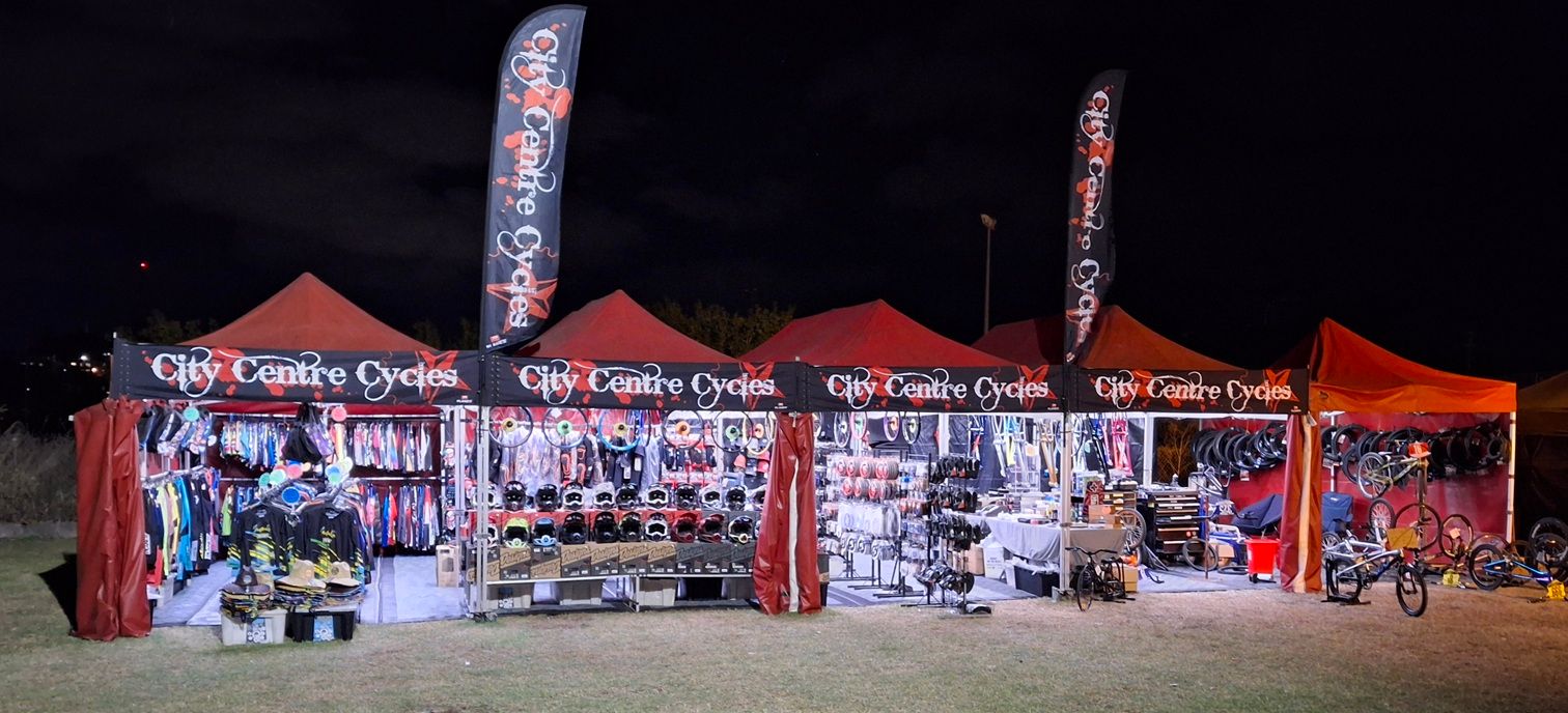 Nighttime outdoor cycling booth lit by red canopy tents, with bikes and gear displayed inside — City Centre Cycles in Berserker, QLD