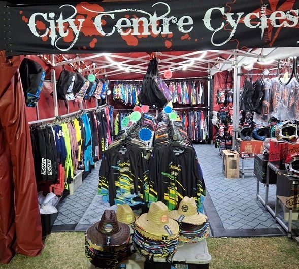 Cycle shop stall with colourful jackets, helmets, and hats under a red canopy titled "City Centre Cycles" — City Centre Cycles in Berserker, QLD