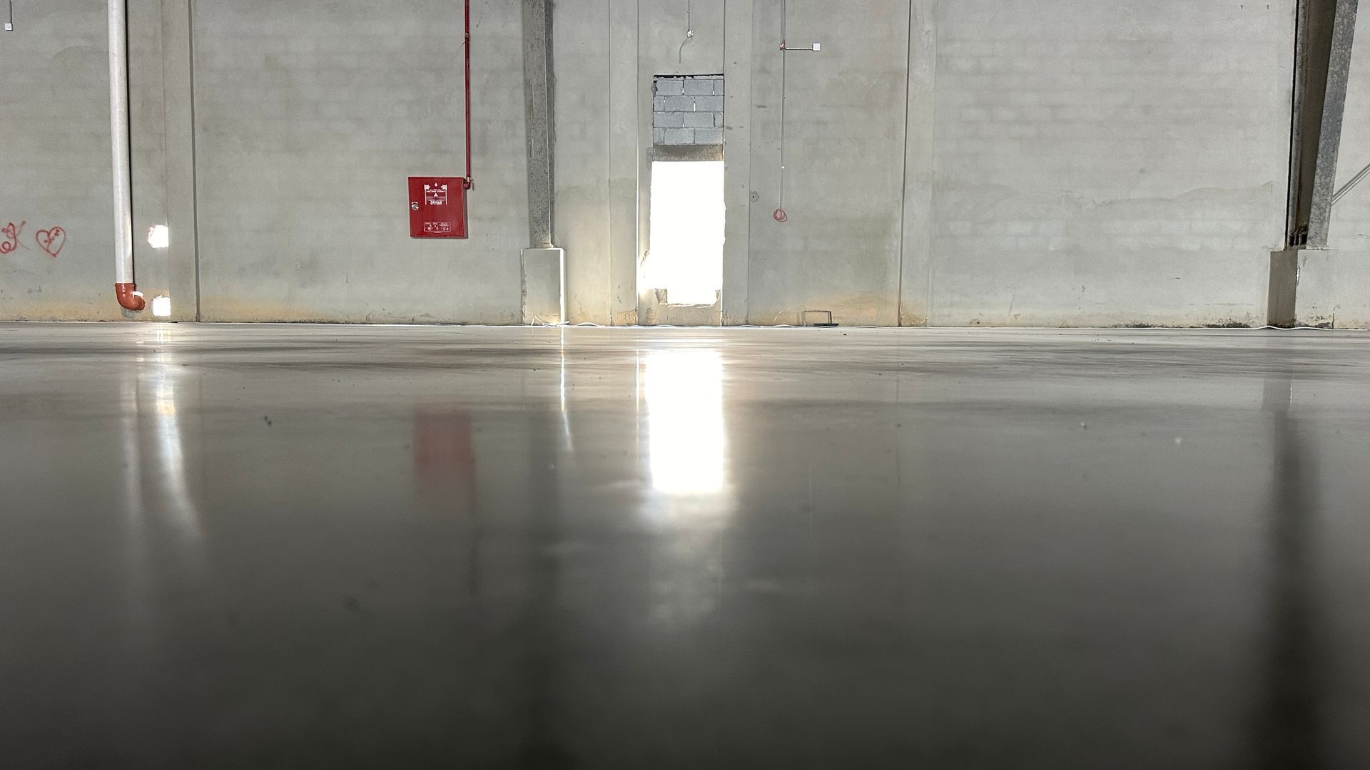 Shiny concrete floor reflecting light in an empty industrial building with bright doorway and fire equipment.