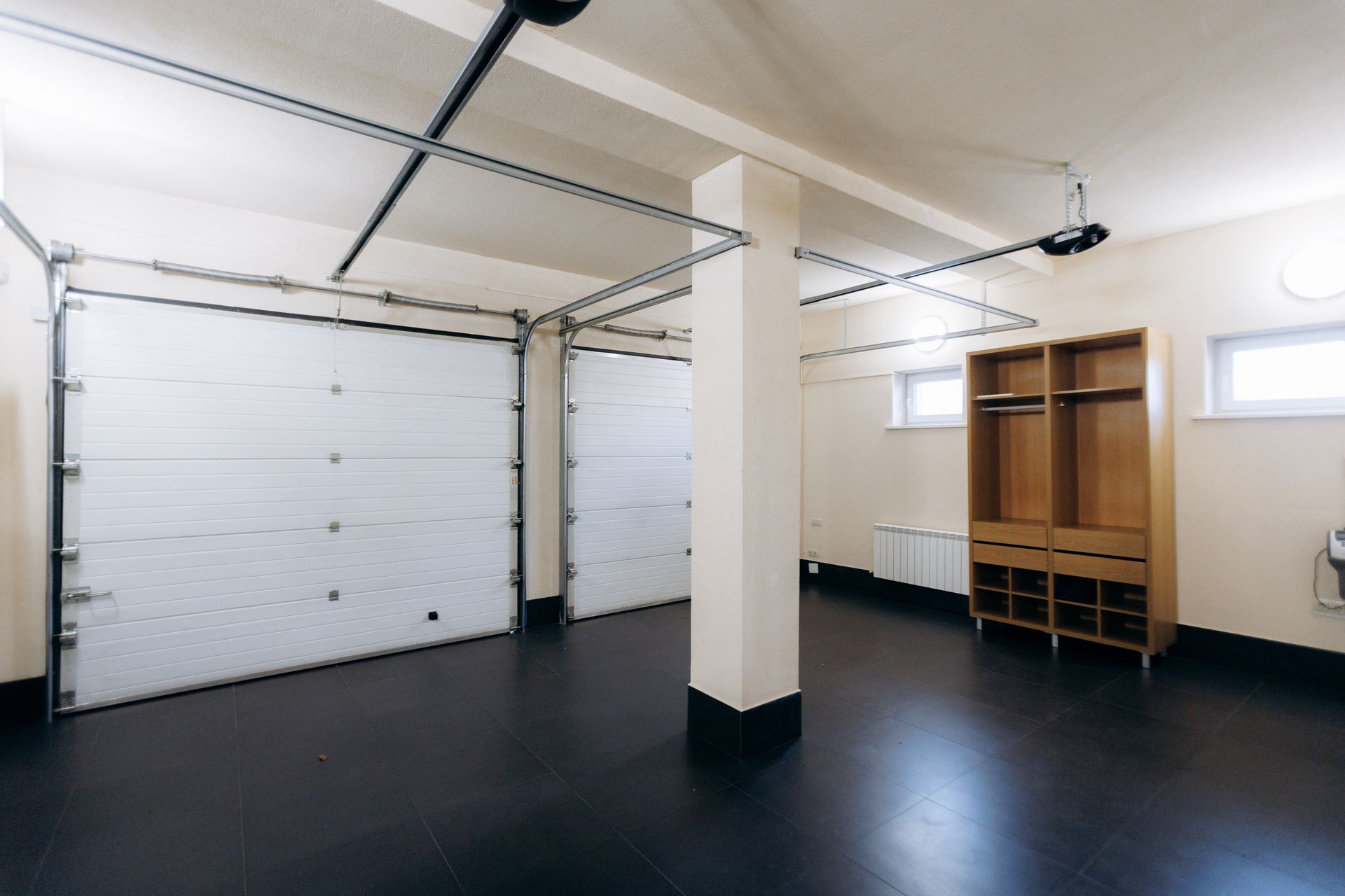 Garage interior with closed white garage doors, brown wooden cabinet, and dark floor. Garage interior with closed white garage doors, brown wooden cabinet, and dark floor.