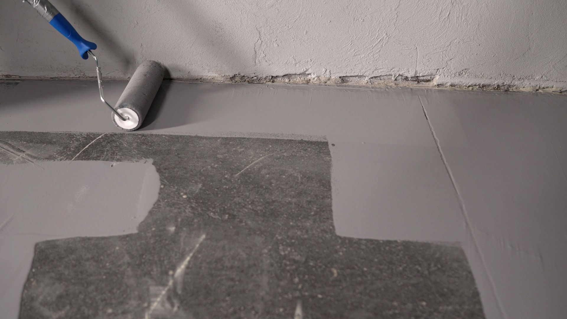 A paint roller applies gray coating to a concrete floor against a gray wall.