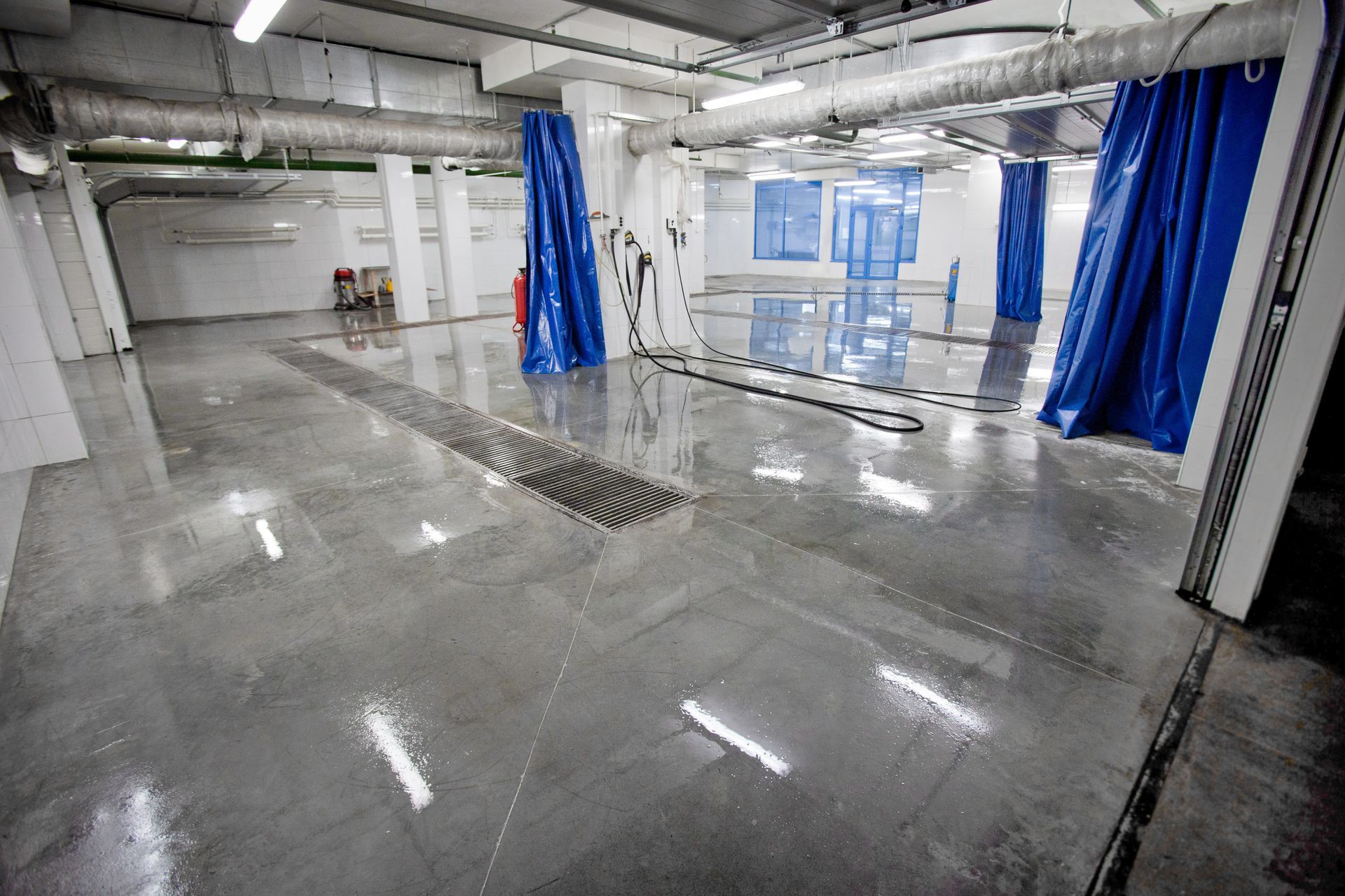 Indoor car wash bay with wet, gray concrete floor, blue curtains, and overhead lighting.