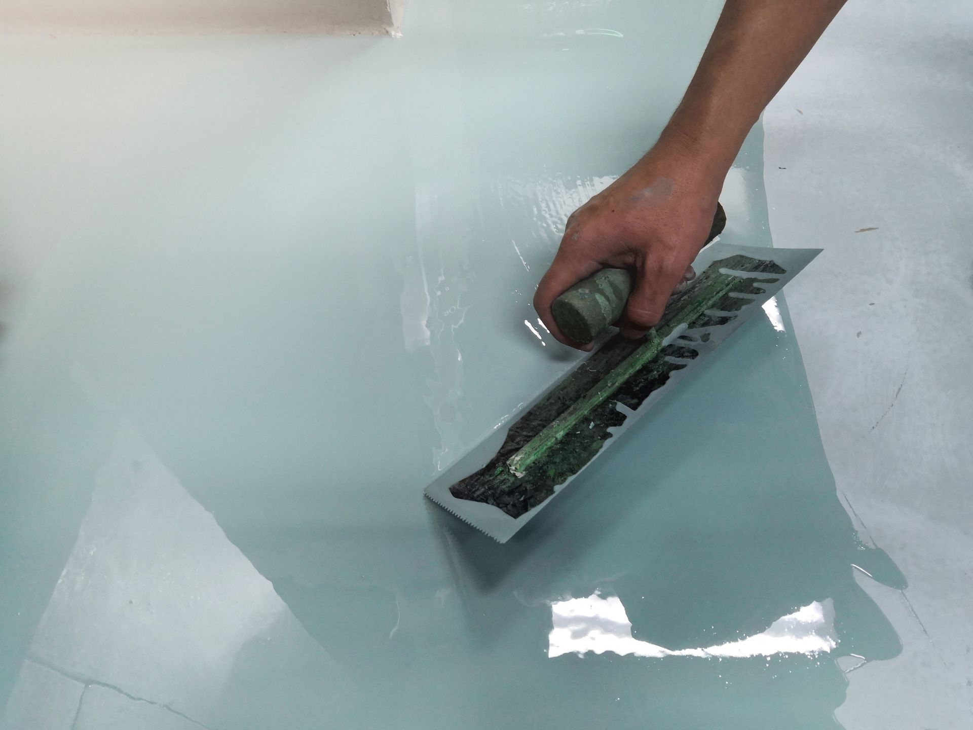 Hand using a trowel to smooth light blue epoxy on a floor.