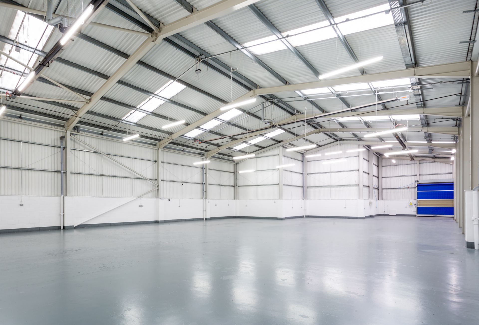 Empty warehouse interior with a gray floor and corrugated metal ceiling, lit by overhead lights and skylights. Empty warehouse interior with a gray floor and corrugated metal ceiling, lit by overhead lights and skylights.