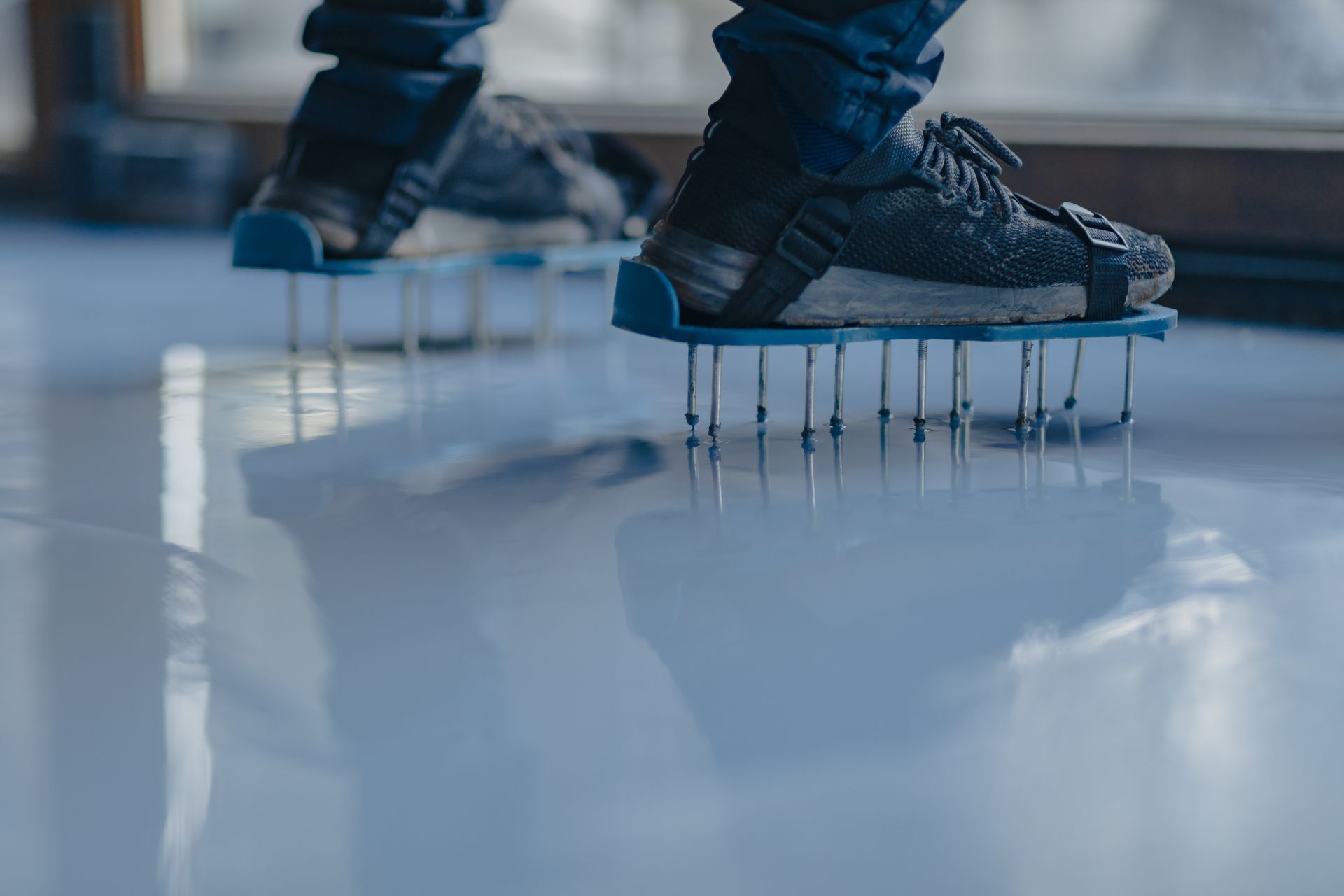 Person's shoes with spikes standing on a reflective surface, likely epoxy. Dark sneakers, blue spikes and holders.
