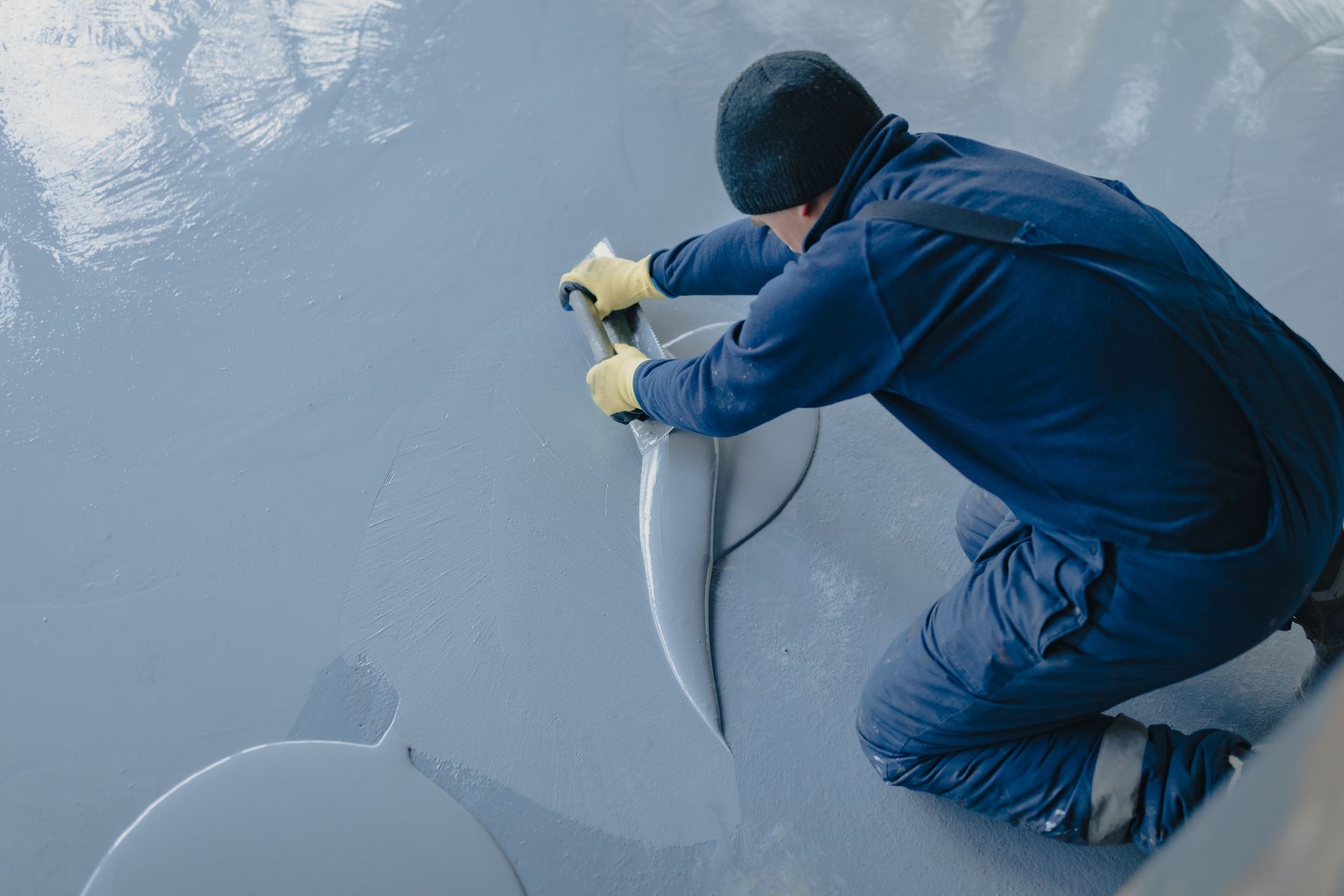 Person in blue coveralls smoothing fresh gray coating on a floor with a tool.