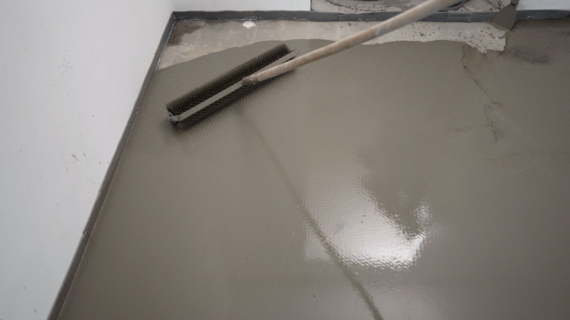 Floor being leveled with a long-handled tool; wet, gray material filling a room's floor space next to a white wall. Floor being leveled with a long-handled tool; wet, gray material filling a room's floor space next to a white wall.