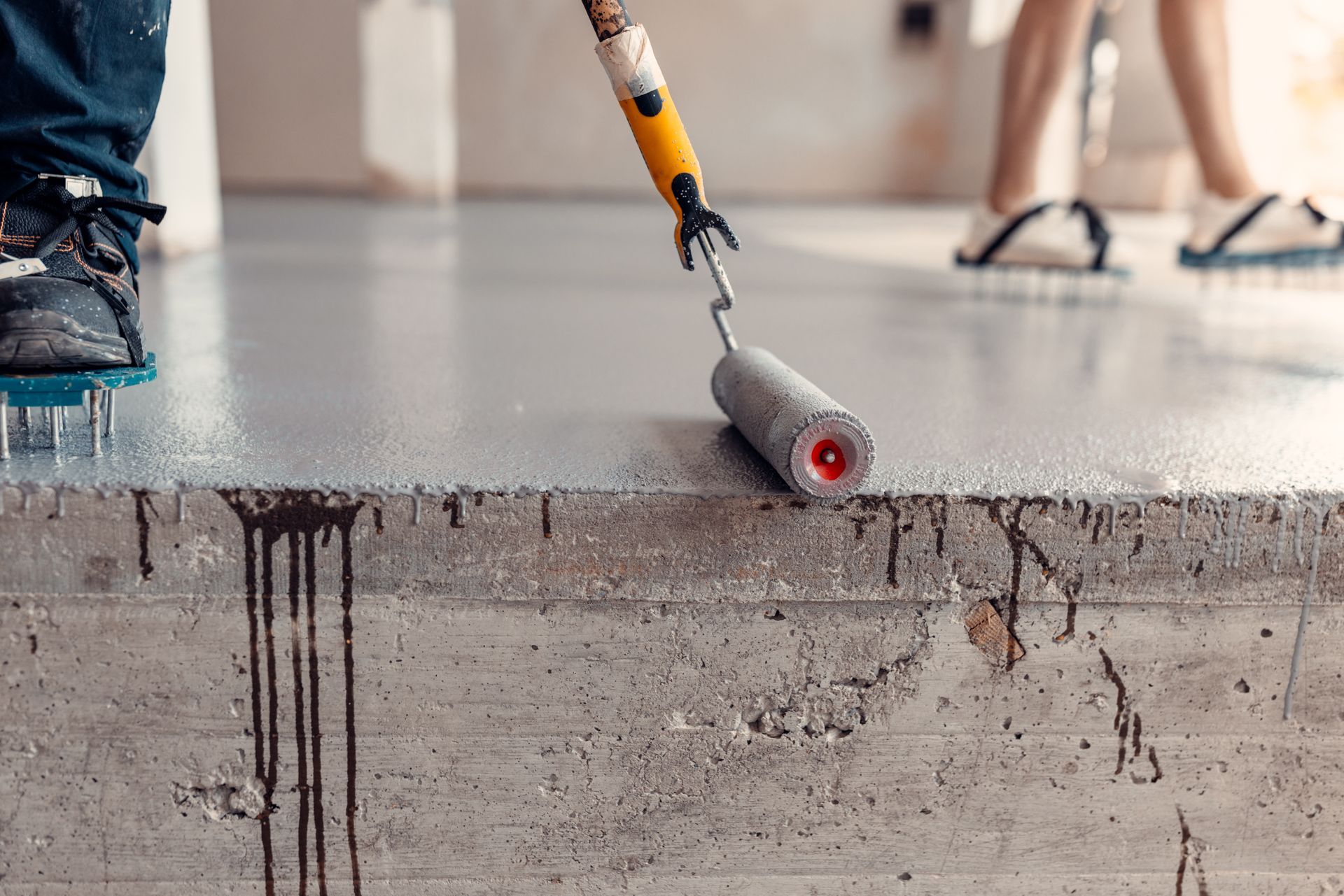 Person using a paint roller on a concrete floor, with paint dripping down the side.