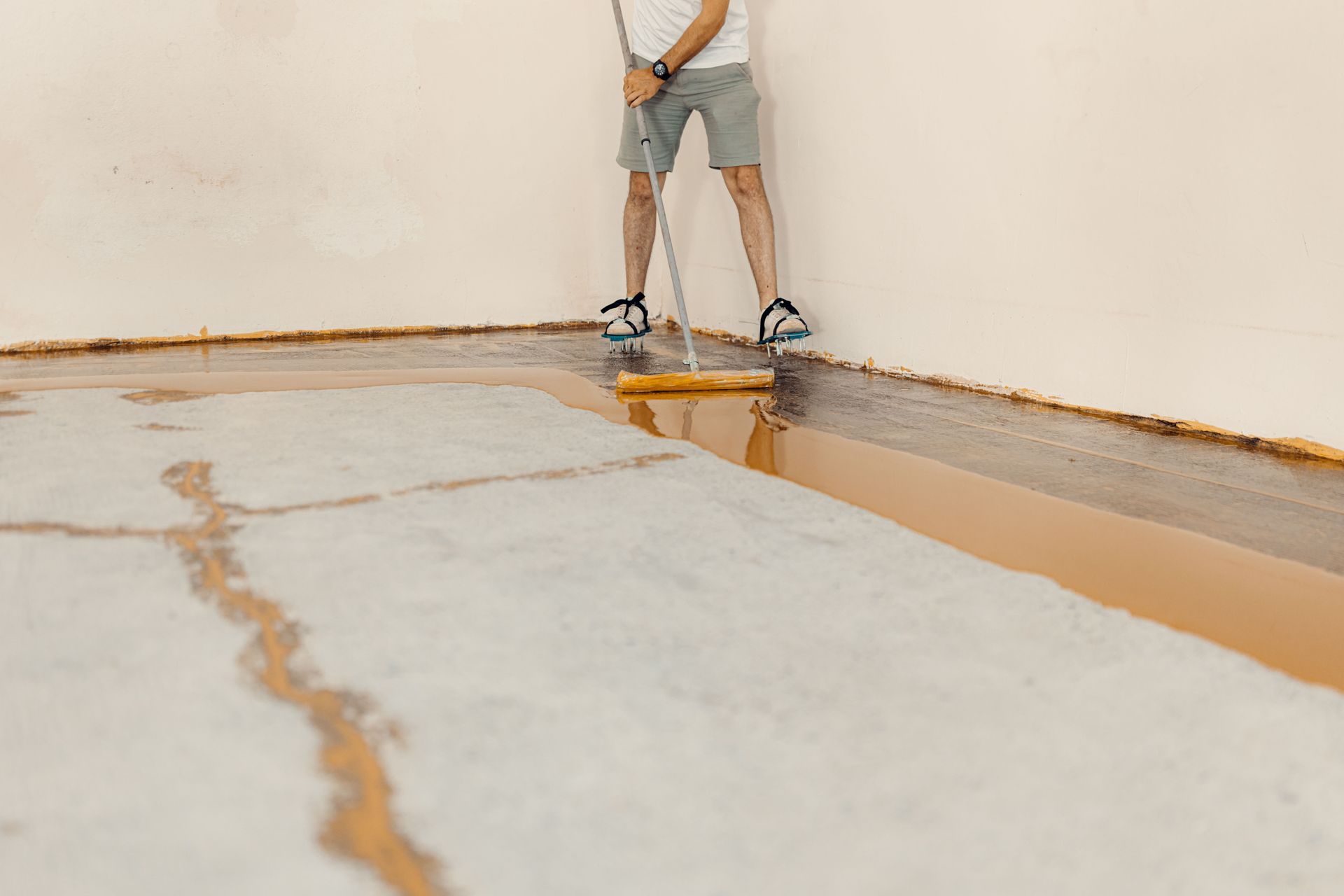 Person applies liquid coating to concrete floor with a roller. Corner of room is visible.