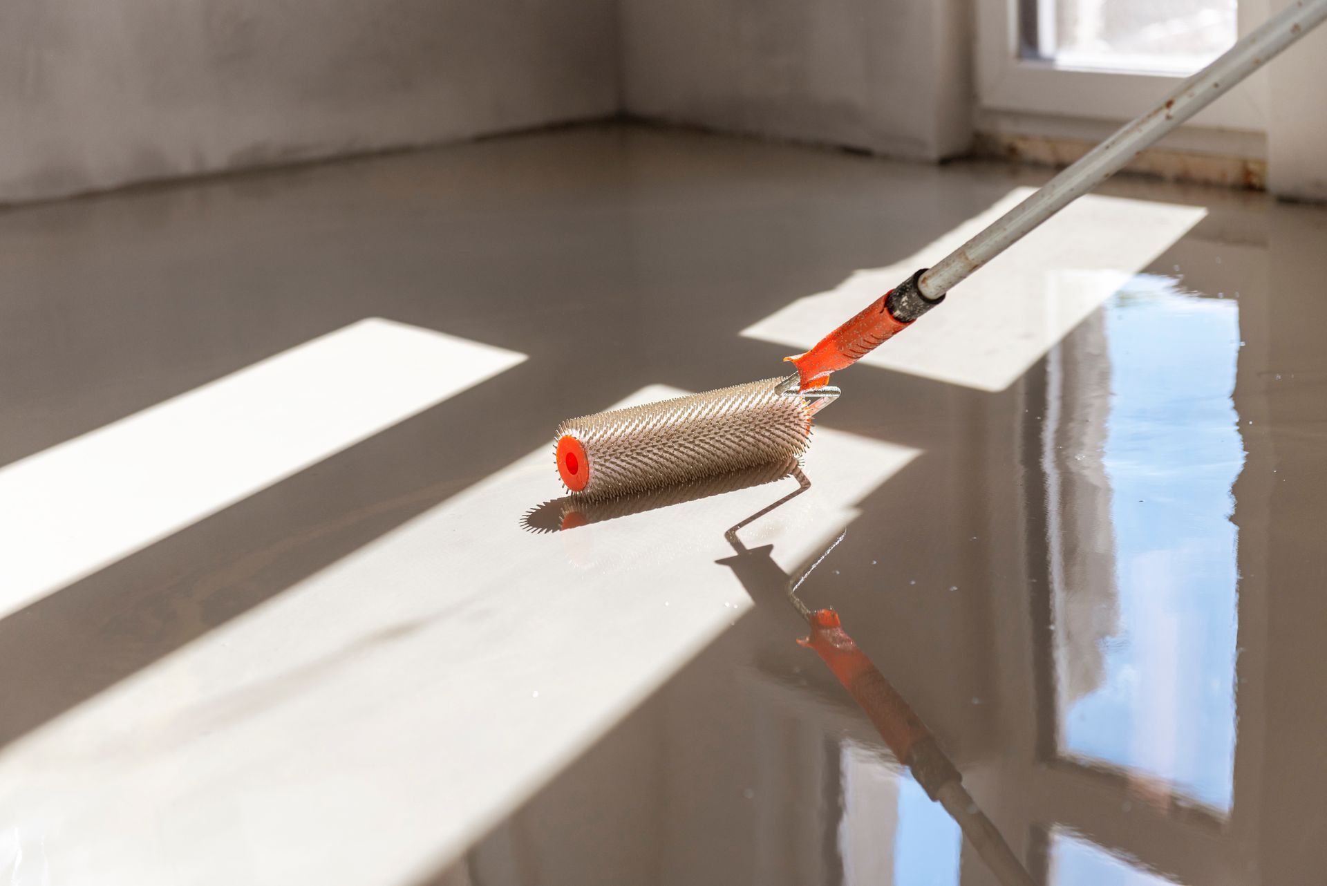 Roller applying glossy, light-colored sealant to a smooth floor in a room with window light. Roller applying glossy, light-colored sealant to a smooth floor in a room with window light.