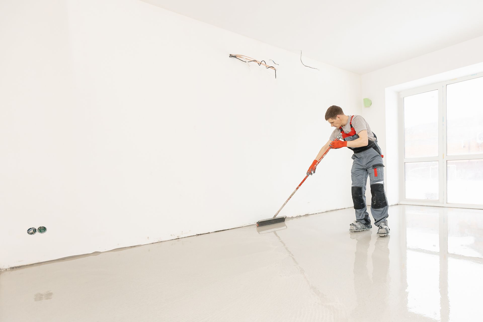 Man smoothing a gray floor with a long-handled tool in a white room with a window. Man smoothing a gray floor with a long-handled tool in a white room with a window.